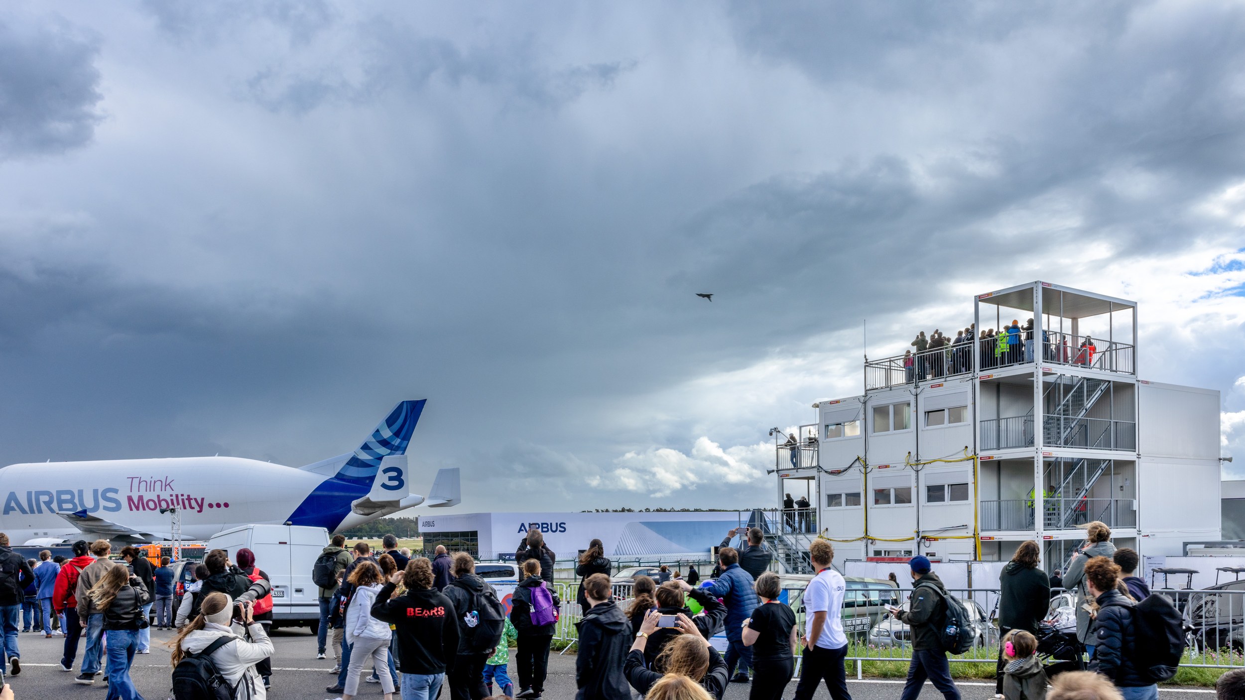 Eine dichte Besuchermenge blickt und fotografiert in den Himmel, wo ein kleiner Kampfjet vor dunklen Wolken fliegt.