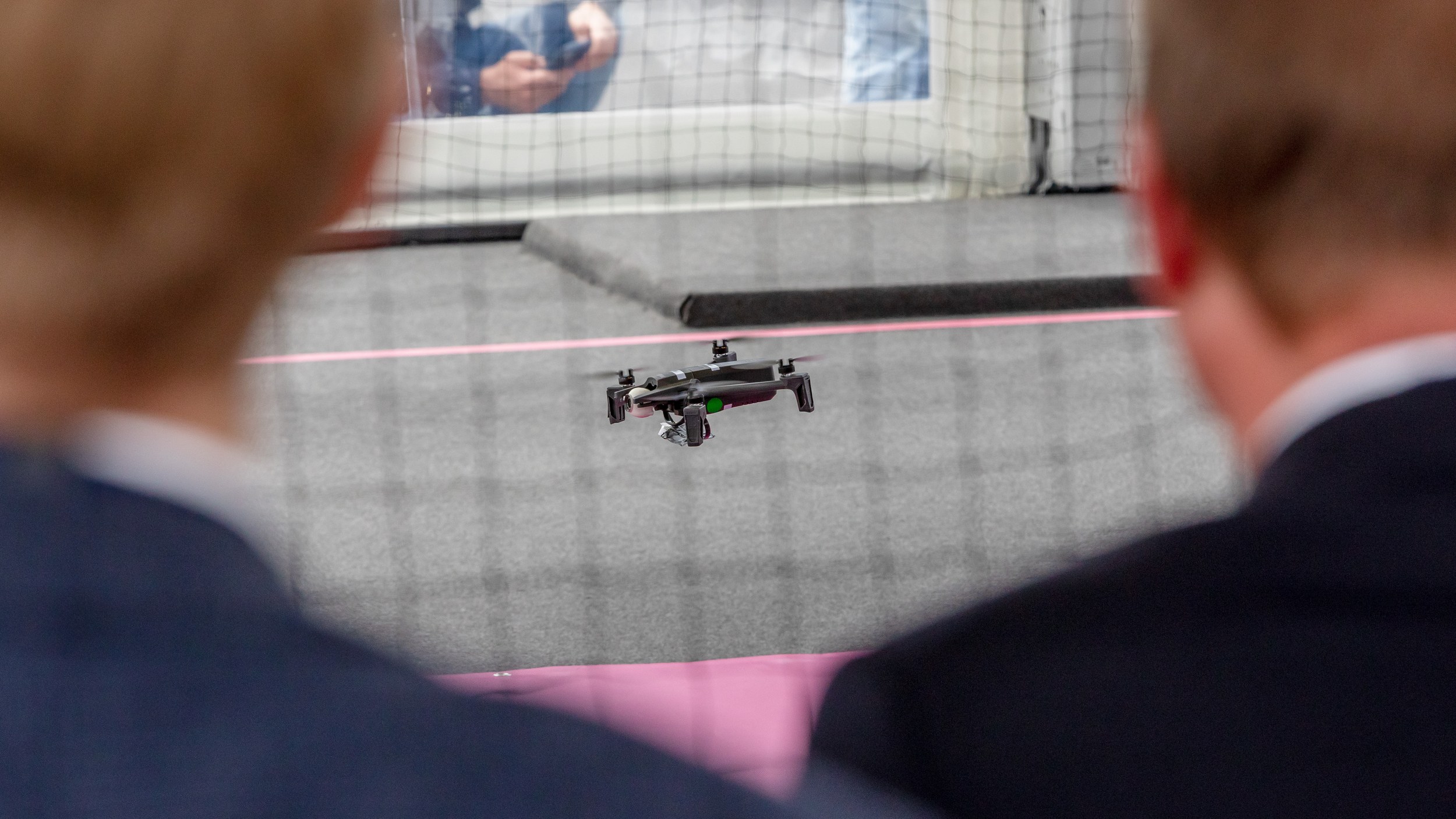 Eine kleine Quadrocopter-Drohne schwebt knapp über dem Boden in einer mit Schutznetz gesicherten Flugzone. Zwei groß und