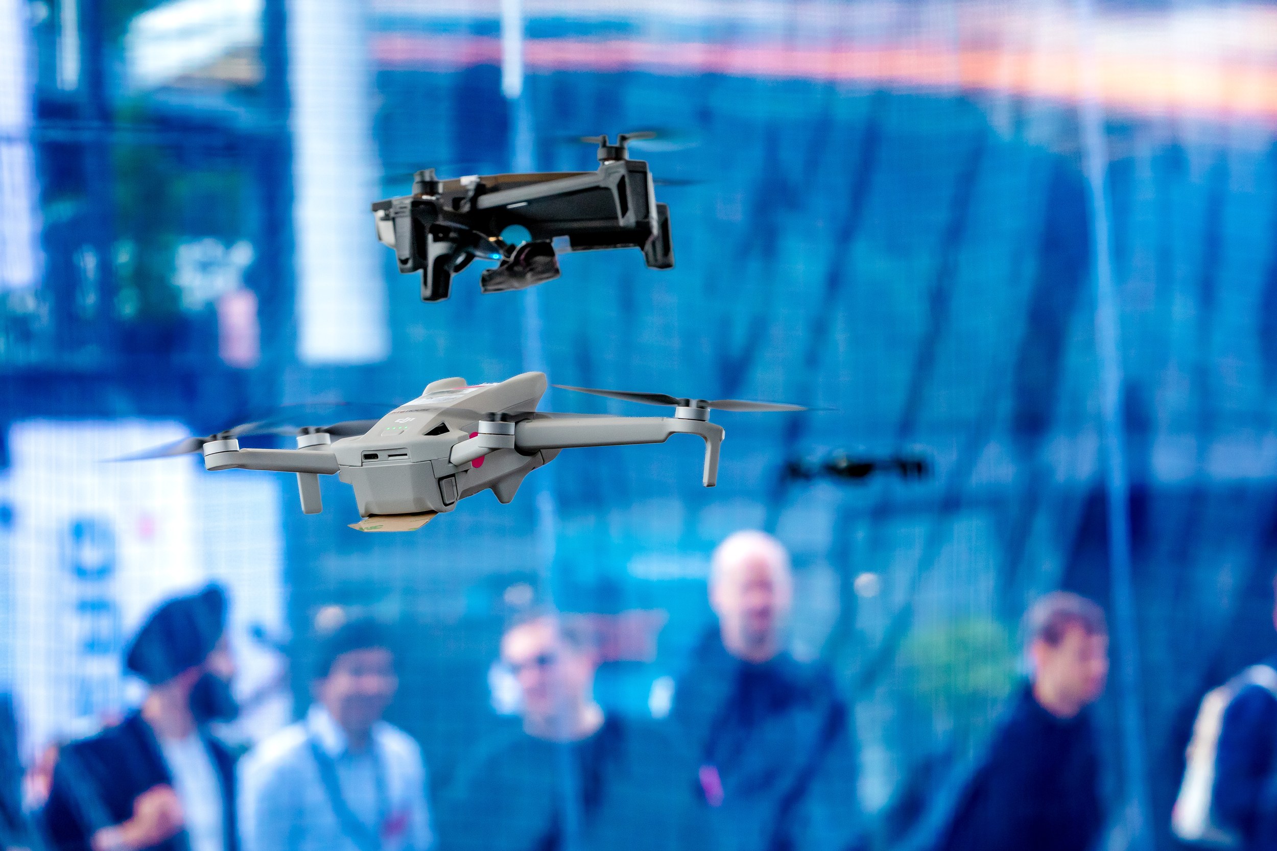Zwei scharf erkennbare Quadrocopter schweben übereinander vor einem blau verglasten Hallenhintergrund, eine graue Drohne
