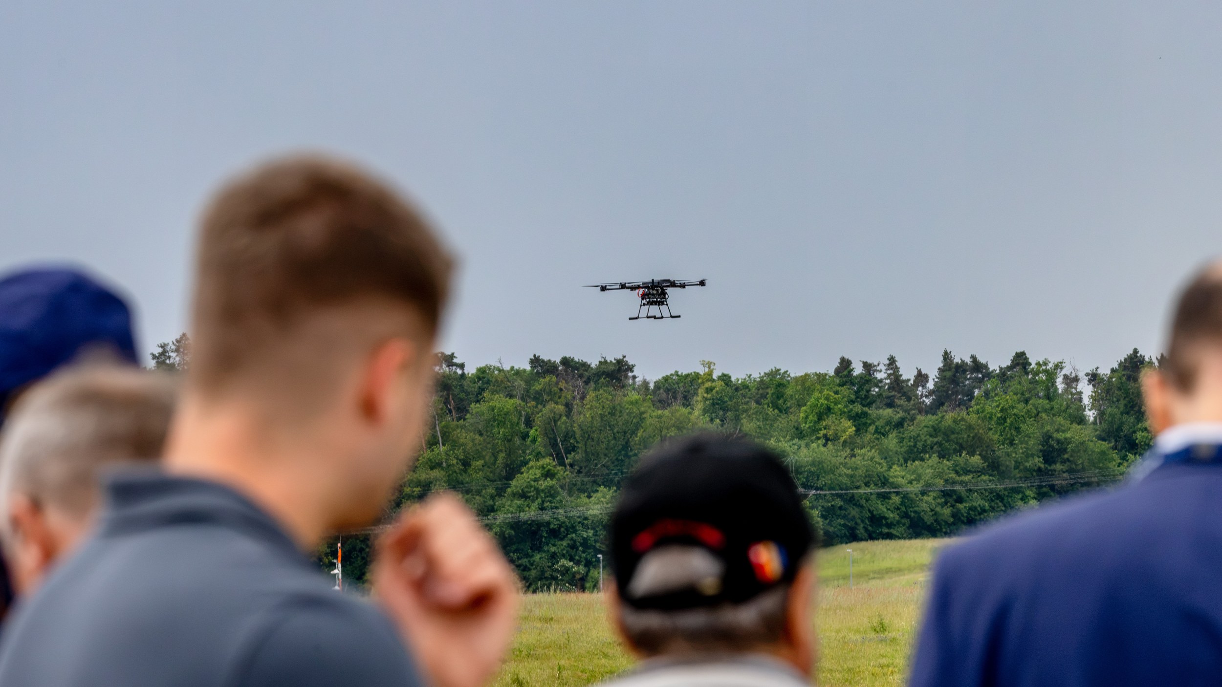 Mehrere unscharf angeschnittene Zuschauer blicken beim Flying Display der ILA Berlin 2026 auf eine Mehrrotordrohne. Das 