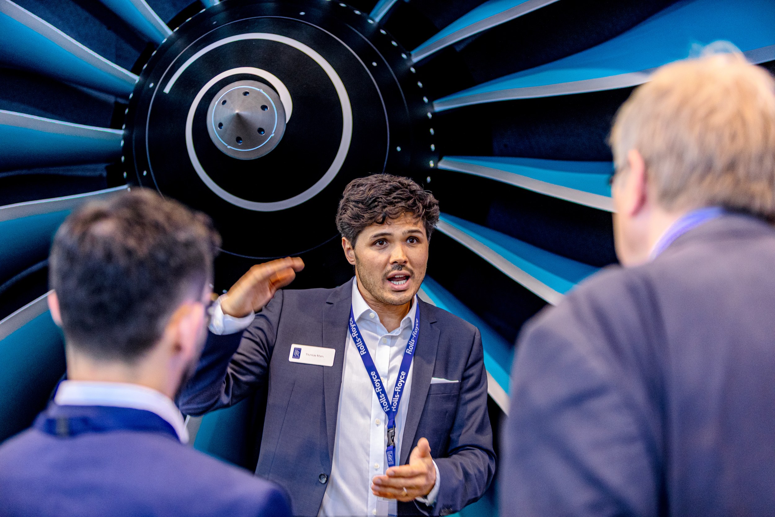 Ein Rolls-Royce-Mitarbeiter erklärt zwei Besuchern vor einem großen blau-schwarzen Turbofan-Display ein technisches …