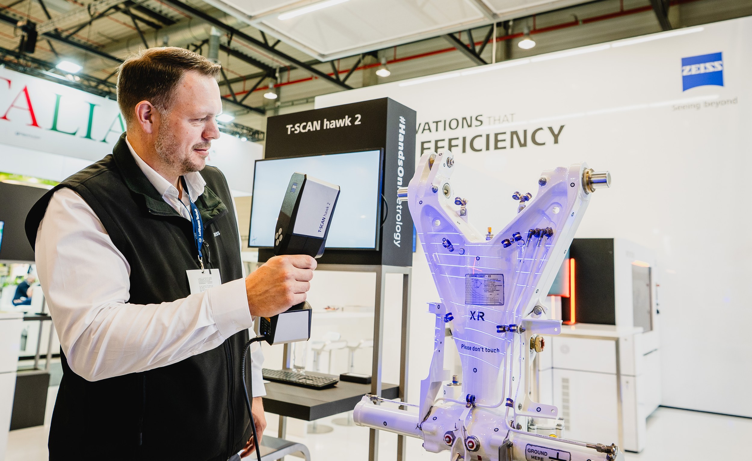 A Zeiss demonstrator points a handheld T-SCAN hawk 2 at a light-colored structural component covered with fittings and tracking markers. A product monitor and ZEISS and HandsOnMetrology branding appear behind the demonstration at a white exhibition booth.