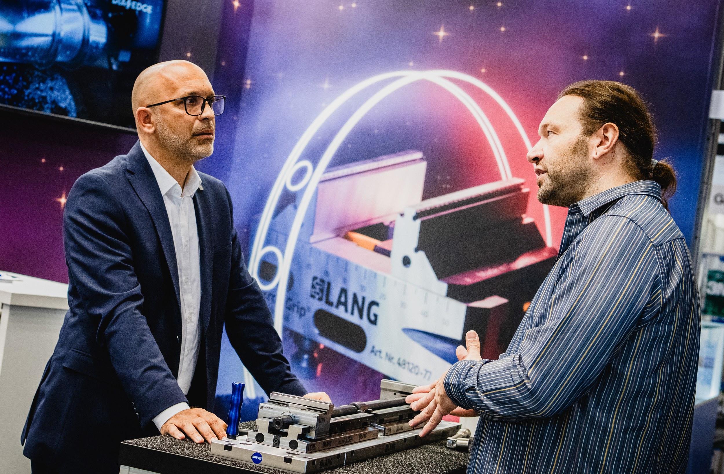 Two men talk at the Lang Technik booth over a clamping system with a metal component mounted on a demonstration table. The man on the right gestures above rails and fixtures while a large product graphic behind them displays LANG and item number 4620-77.