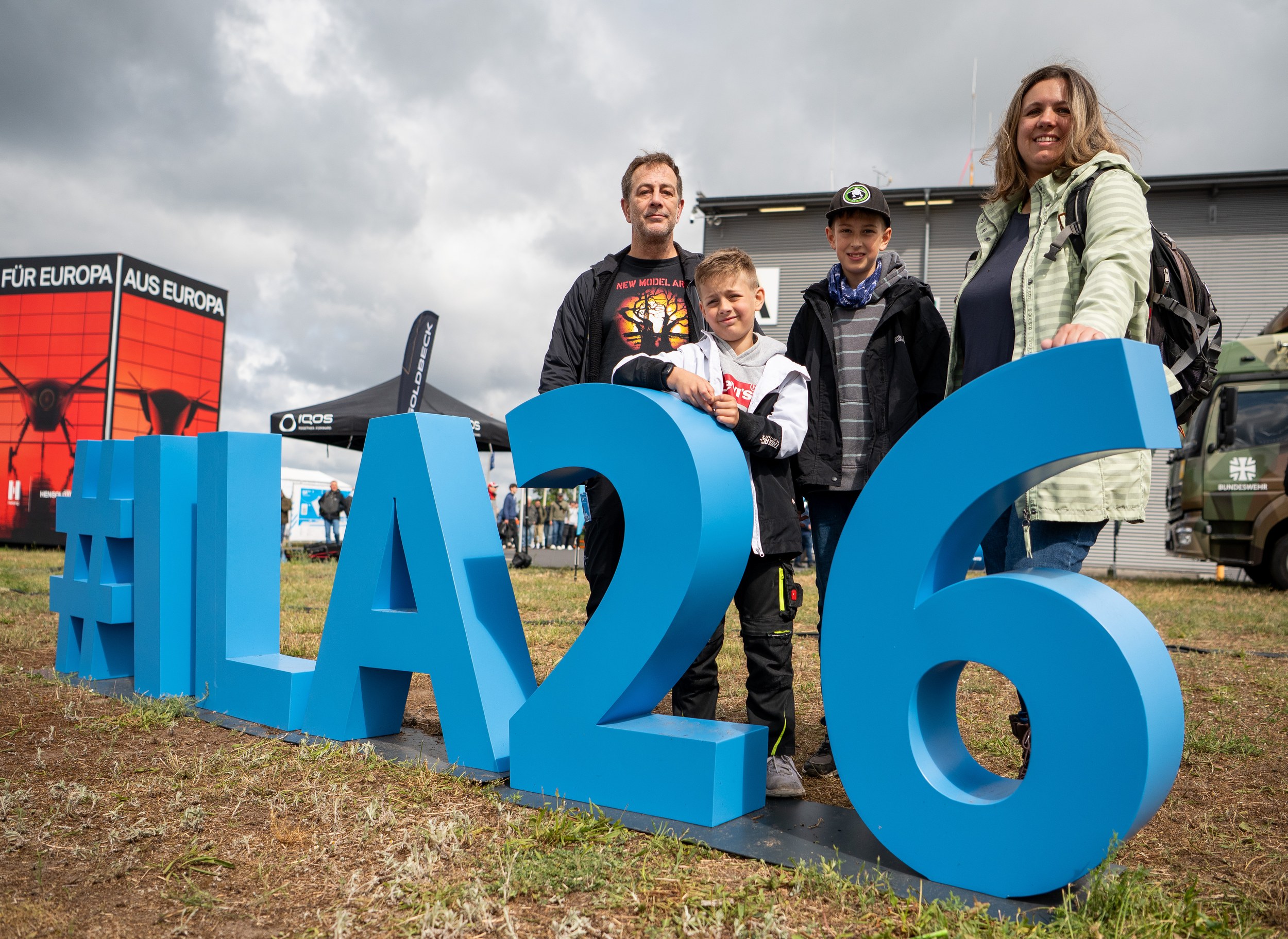 Zwei Erwachsene und zwei Kinder posieren im Freigelände hinter den großen blauen #ILA26-Buchstaben für ein …