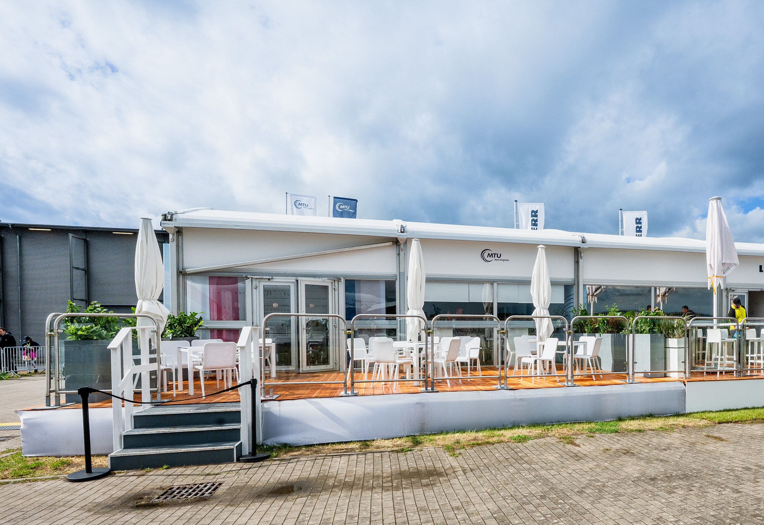 Chalets at ILA 2026 seen from the terrace side: white modules with a glass front and double glass door, terrace with glass railing and stairs.