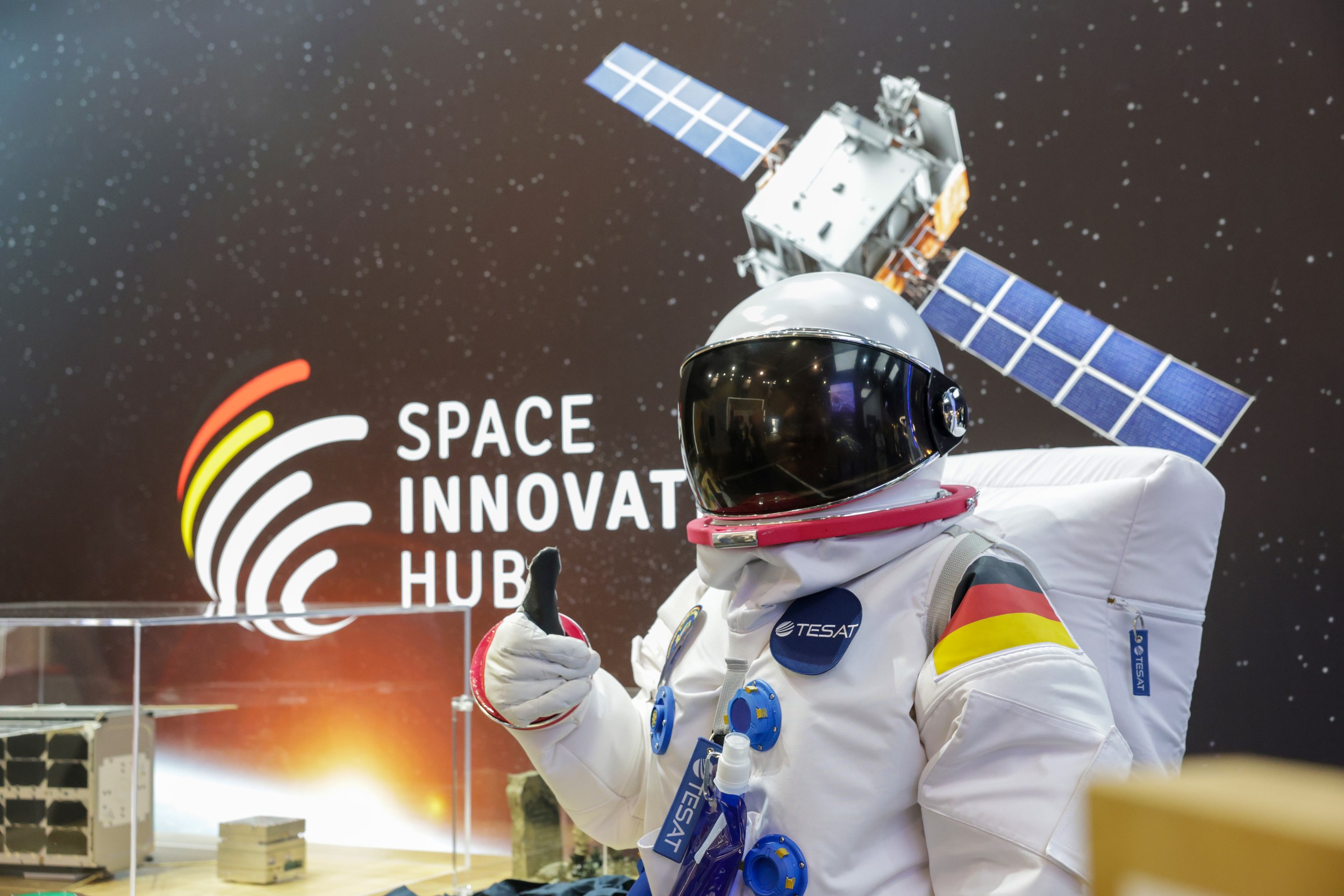 A person in a white astronaut costume with a TESAT patch, a German flag on the arm and a mirrored visor gives a thumbs u