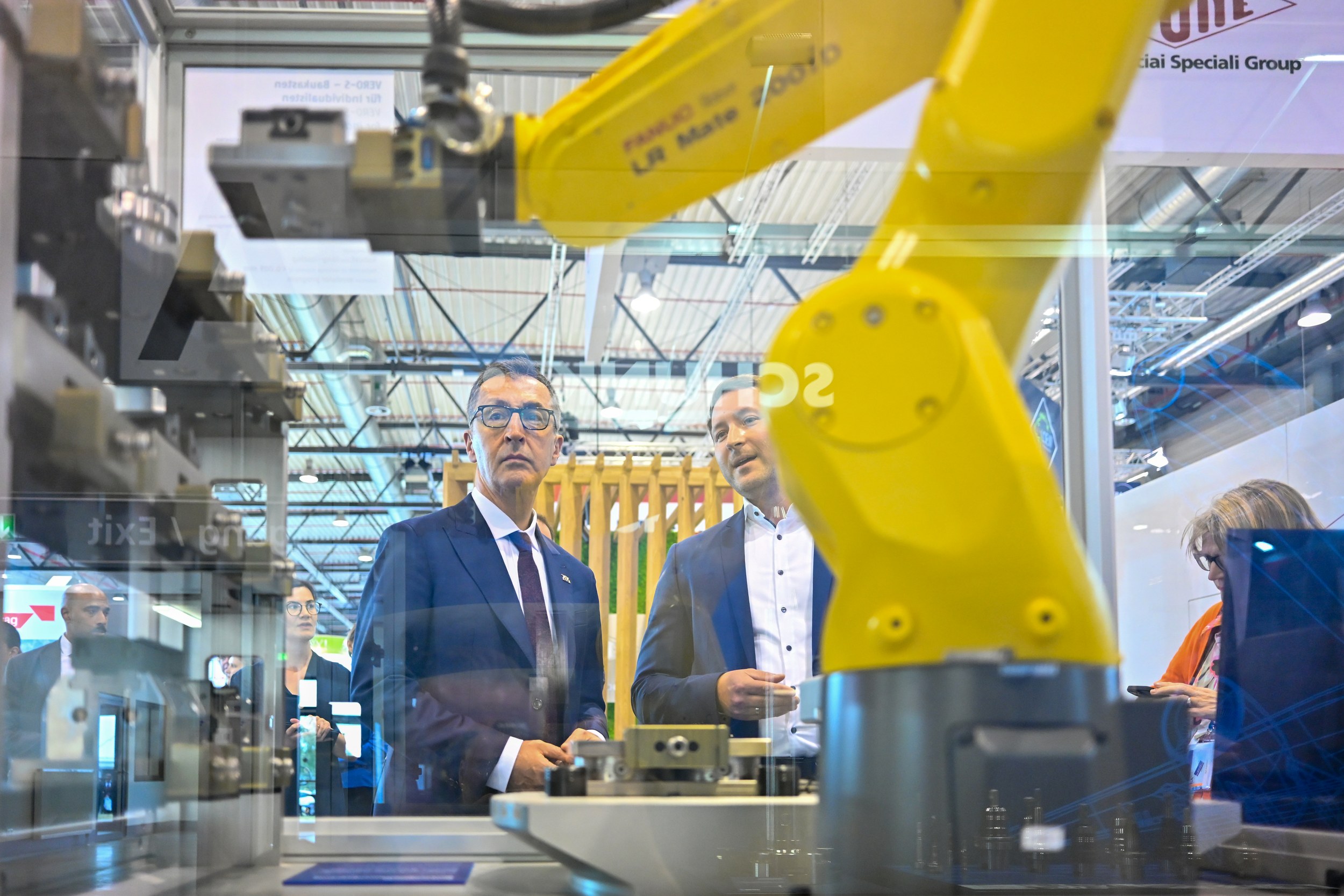 Cem Özdemir looks through a glass panel at a manufacturing cell dominated by a yellow industrial robot in the foreground. A booth representative explains the installation with metal clamping and machining components as the exhibition hall is reflected in the glass.