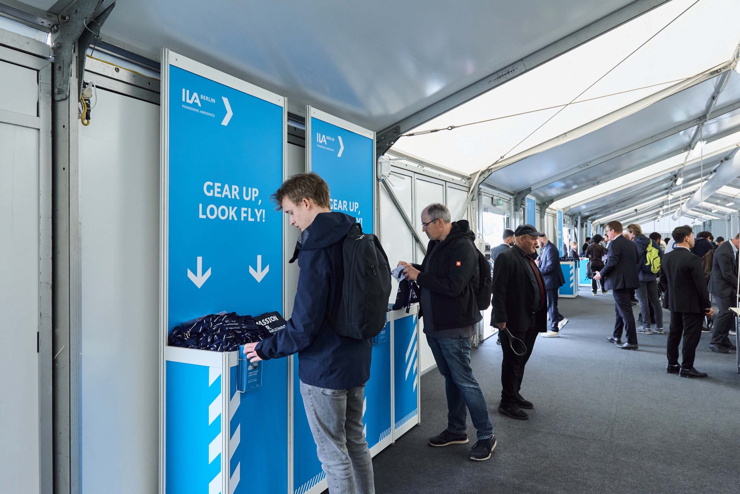 Besucher nehmen gebrandete Lanyards von den blauen Gear-up-look-fly-Ausgabestationen am Eingang der ILA 2026.