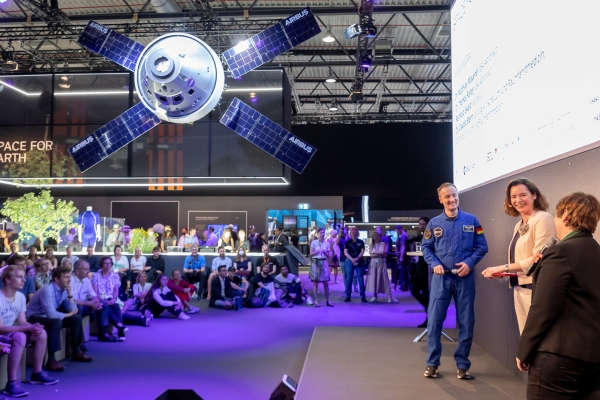 Ein Astronaut und zwei Frauen sprechen auf einer Bühne beim Airbus-Messestand, im Hintergrund ein hängendes Satellitenmodell und Publikum.